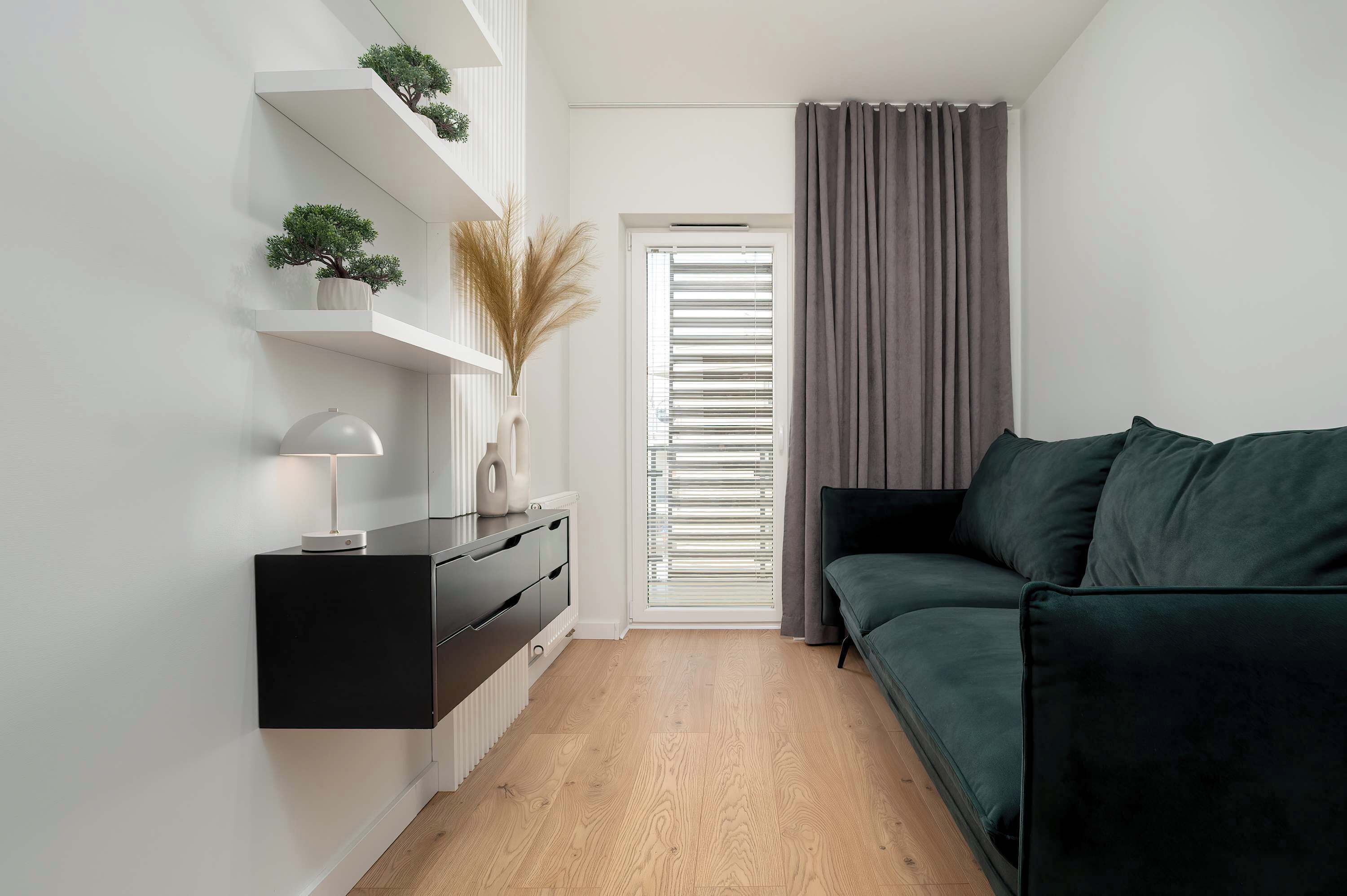 Een moderne woonkamer met een donkergroene bank, een zwarte console tafel met een lamp, en een raam met jaloezieën.