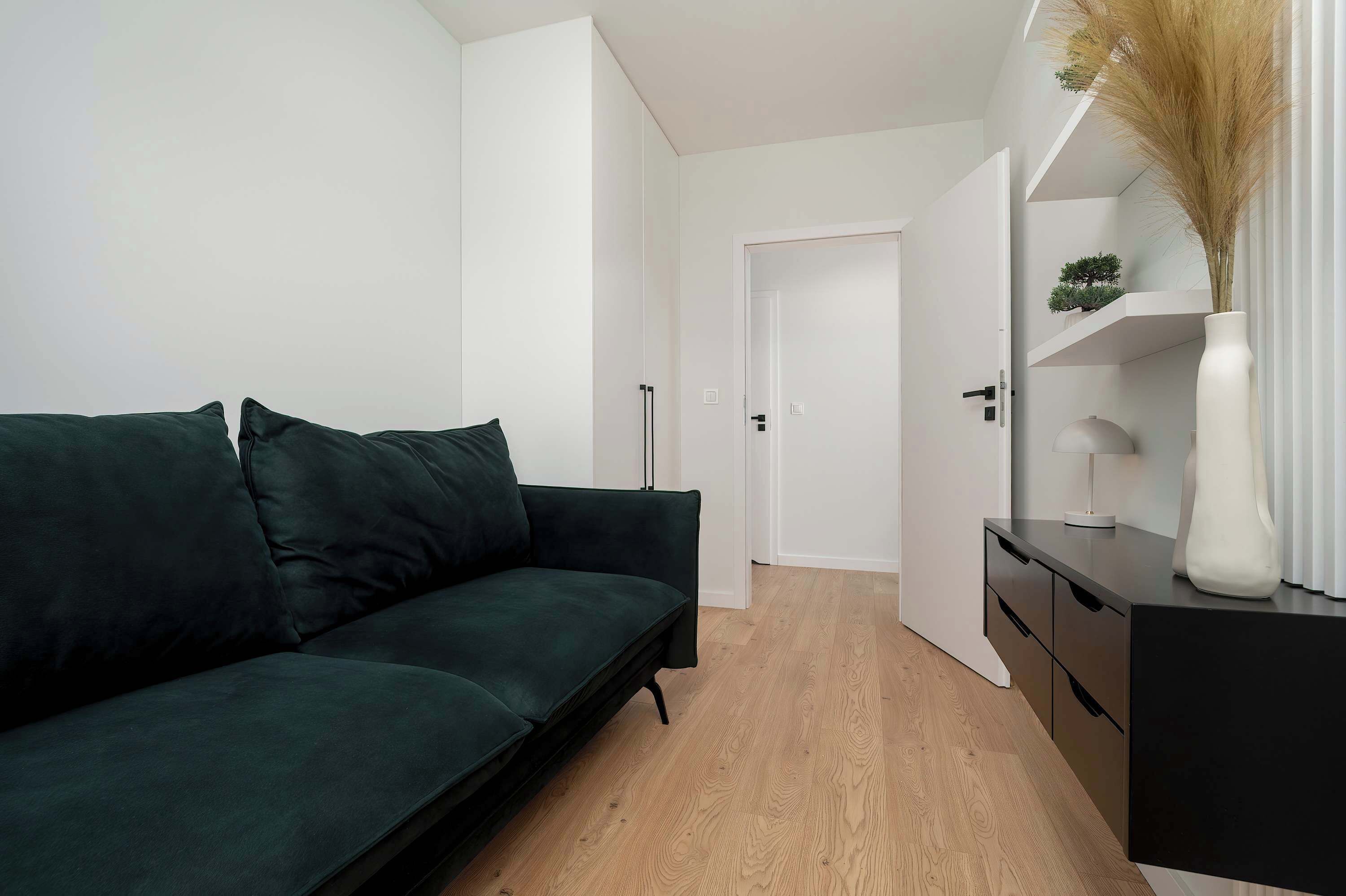 En moderne stue med en mørkegrøn sofa, trægulv, et sort skab og en hvid vase med tørrede planter.
