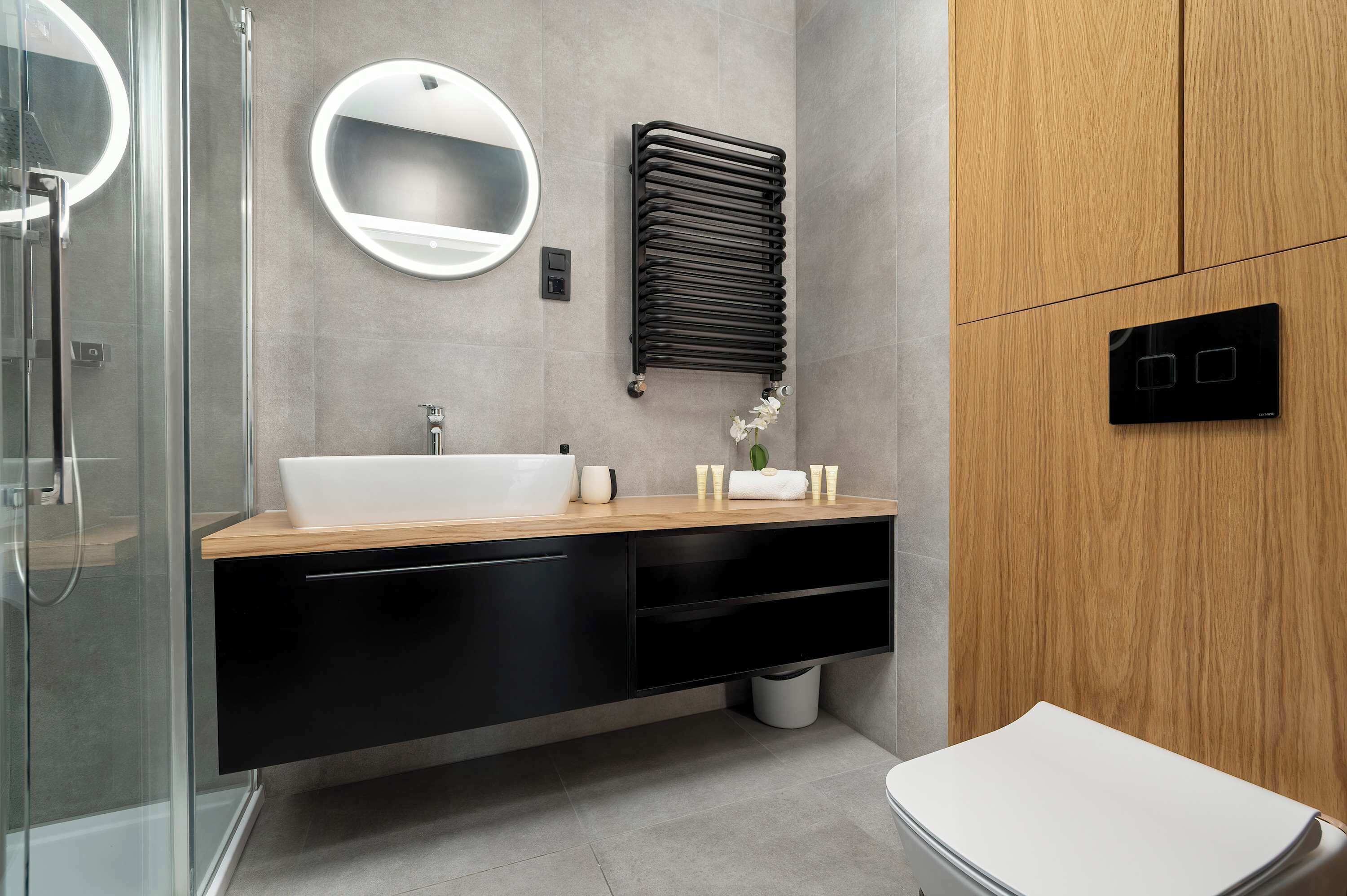 A modern bathroom with a glass shower enclosure, a white sink, a wooden vanity, a round mirror, a towel rack, and a toilet.