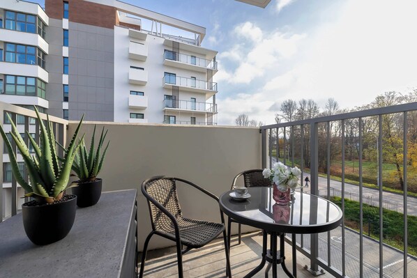 A balcony with two potted aloe plants, a wicker chair, a small round table with a vase of flowers, and a view of a road and trees.