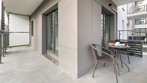 A modern balcony with a metal table and wicker chairs, a glass door, and a view of the building's interior.