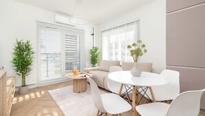 A modern living room with a round dining table, white chairs, a beige sofa, and a coffee table with a plant and a glass of orange juice.