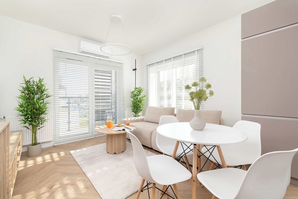 A modern living room with a round dining table, white chairs, a beige sofa, and a coffee table with a plant and a glass of orange juice.