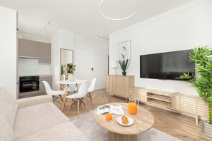 A modern living room with a sofa, dining table, and a TV. There's a kitchen area with cabinets and a microwave.