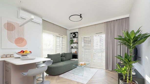 Un salon moderne avec un canapé gris, une table basse en verre, un bureau blanc sur lequel se trouve un saladier rempli de fruits, et une plante en pot.