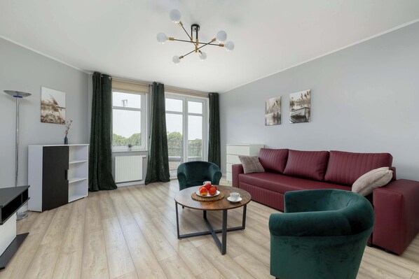 A modern living room with a red sofa, green armchairs, a coffee table with fruit, and a view through large windows.