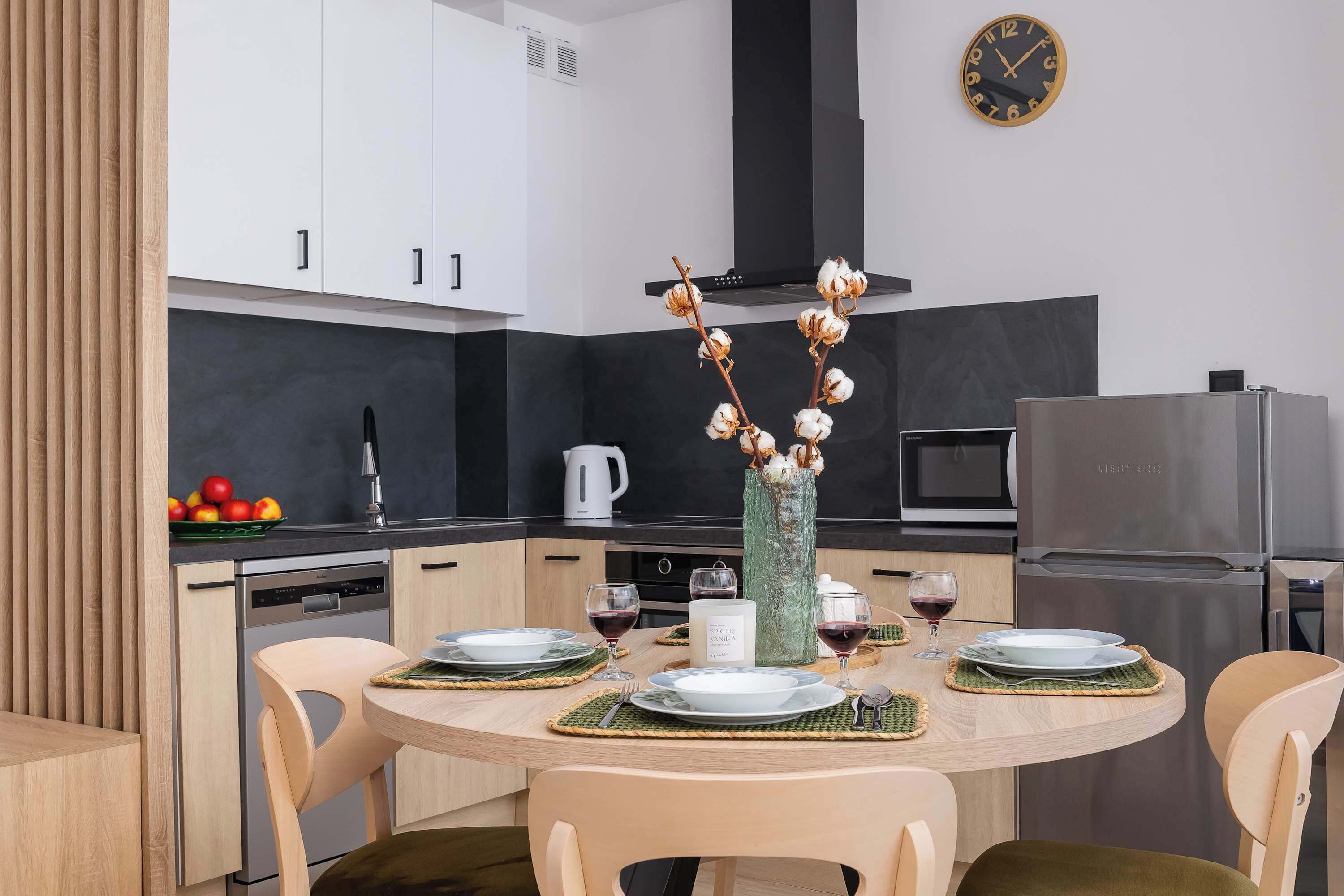 Una cocina moderna con armarios blancos, un refrigerador de acero inoxidable y una mesa de comedor para cuatro personas, con individuales y un jarrón de flores secas.