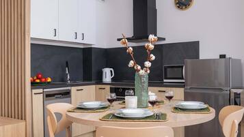 Una cocina moderna con armarios blancos, un refrigerador de acero inoxidable y una mesa de comedor para cuatro personas, con individuales y un jarrón de flores secas.