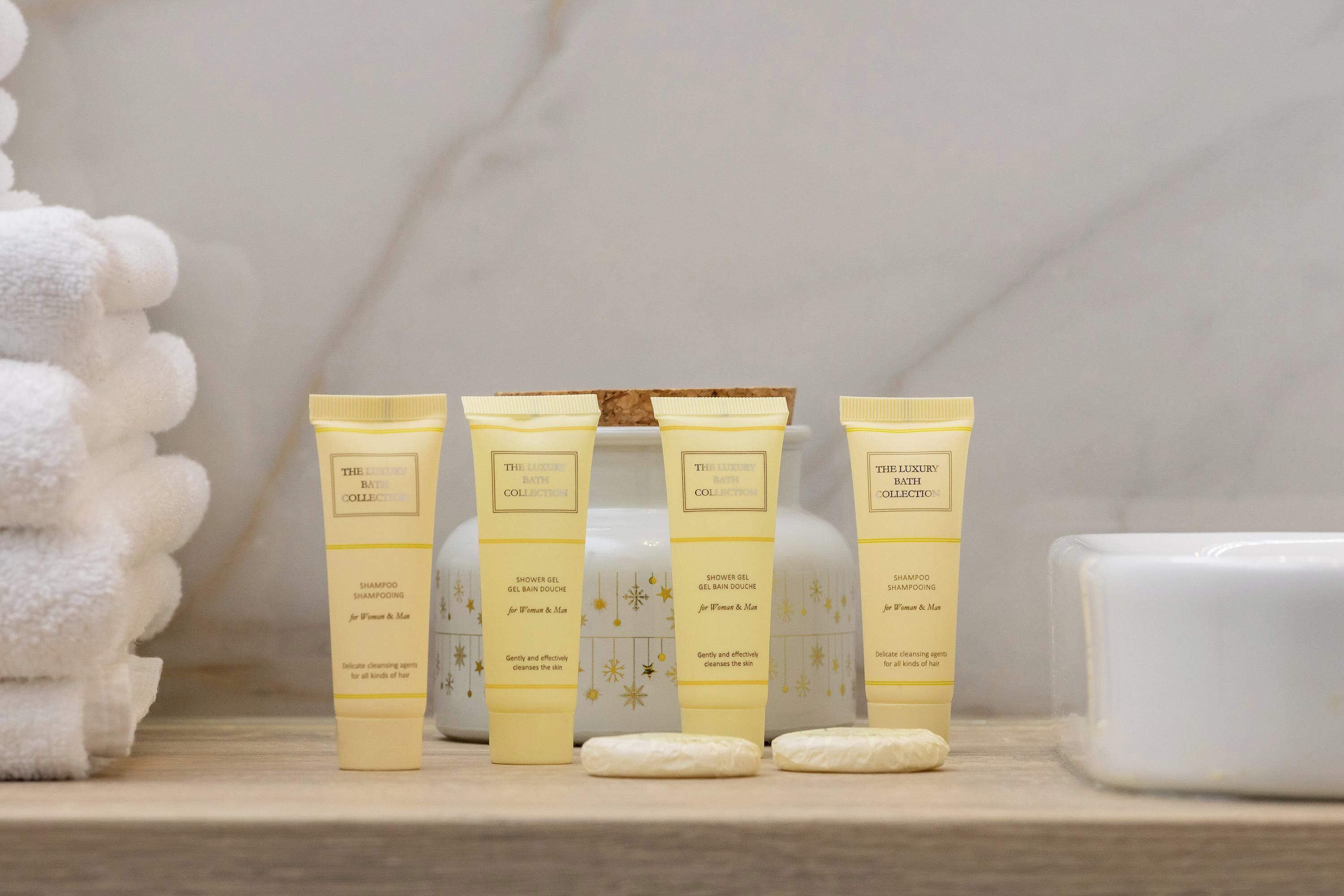 Four yellow tubes of The Luxury Bath Collection on a marble surface with a white candle and a stack of white towels.