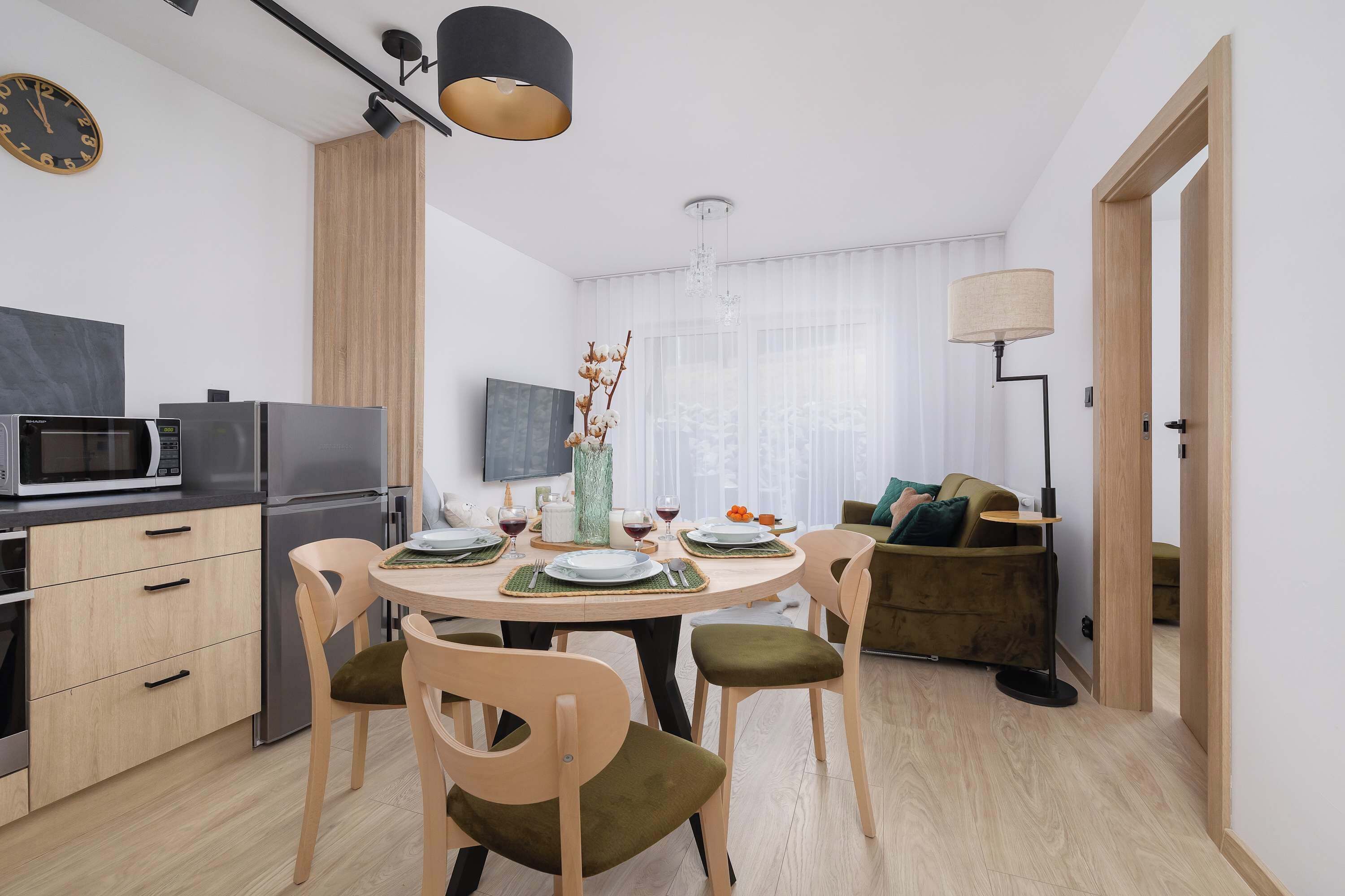 Une cuisine moderne équipée d’un four à micro-ondes, d’un réfrigérateur et de meubles en bois. On y trouve une table à manger avec des chaises et un vase de fleurs. Un canapé et une lampe sont visibles au fond de la pièce.