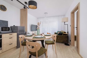 Een moderne keuken met een magnetron, koelkast en houten keukenkastjes. Er staat een eettafel met stoelen en een vaas met bloemen. Achtergrond toont een bank en een lamp.