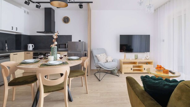 Una cocina moderna con armarios blancos, una zona de comedor con una mesa redonda y sillas, un sillón gris y un televisor en la sala de estar.