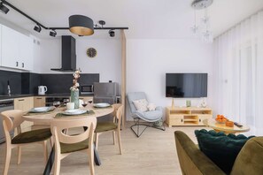 Una cocina moderna con armarios blancos, una zona de comedor con una mesa redonda y sillas, un sillón gris y un televisor en la sala de estar.