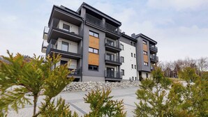Un edificio residencial de varios pisos con balcones, un muro de contención de piedra y una zona pavimentada con árboles.