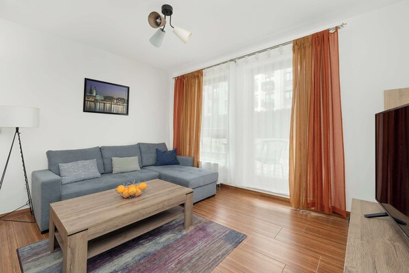 A modern living room with a grey sectional sofa, a wooden coffee table with fruit, a floor lamp, and a large window with sheer curtains.