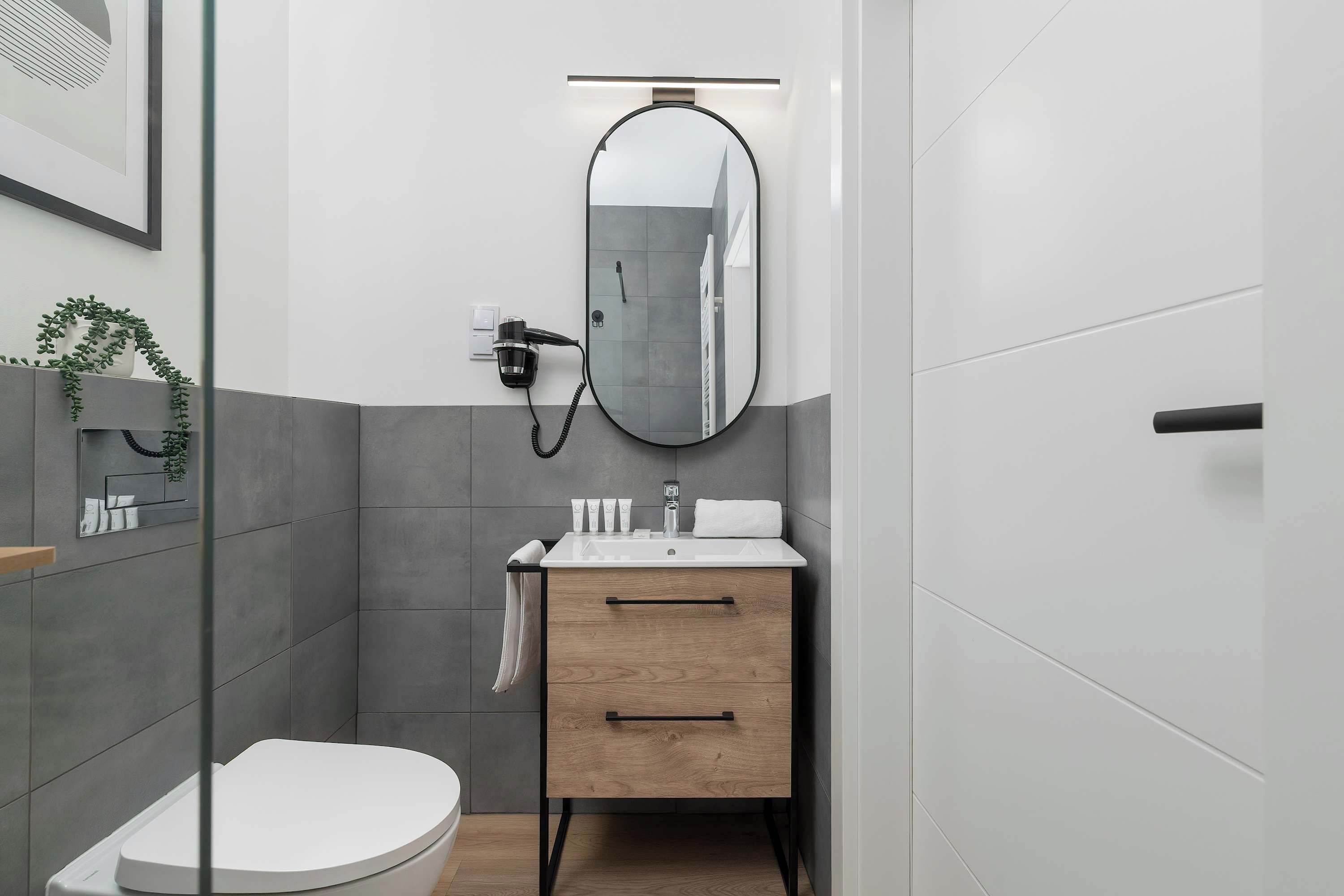 A modern bathroom with a minimalist design, featuring gray tiles, a white toilet, a wooden vanity with black handles, and an oval mirror, exudes elegance and functionality.