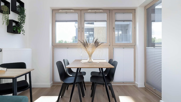 Un comedor moderno con una mesa de madera y sillas negras, un jarrón con plantas secas y ventanas amplias con persianas.