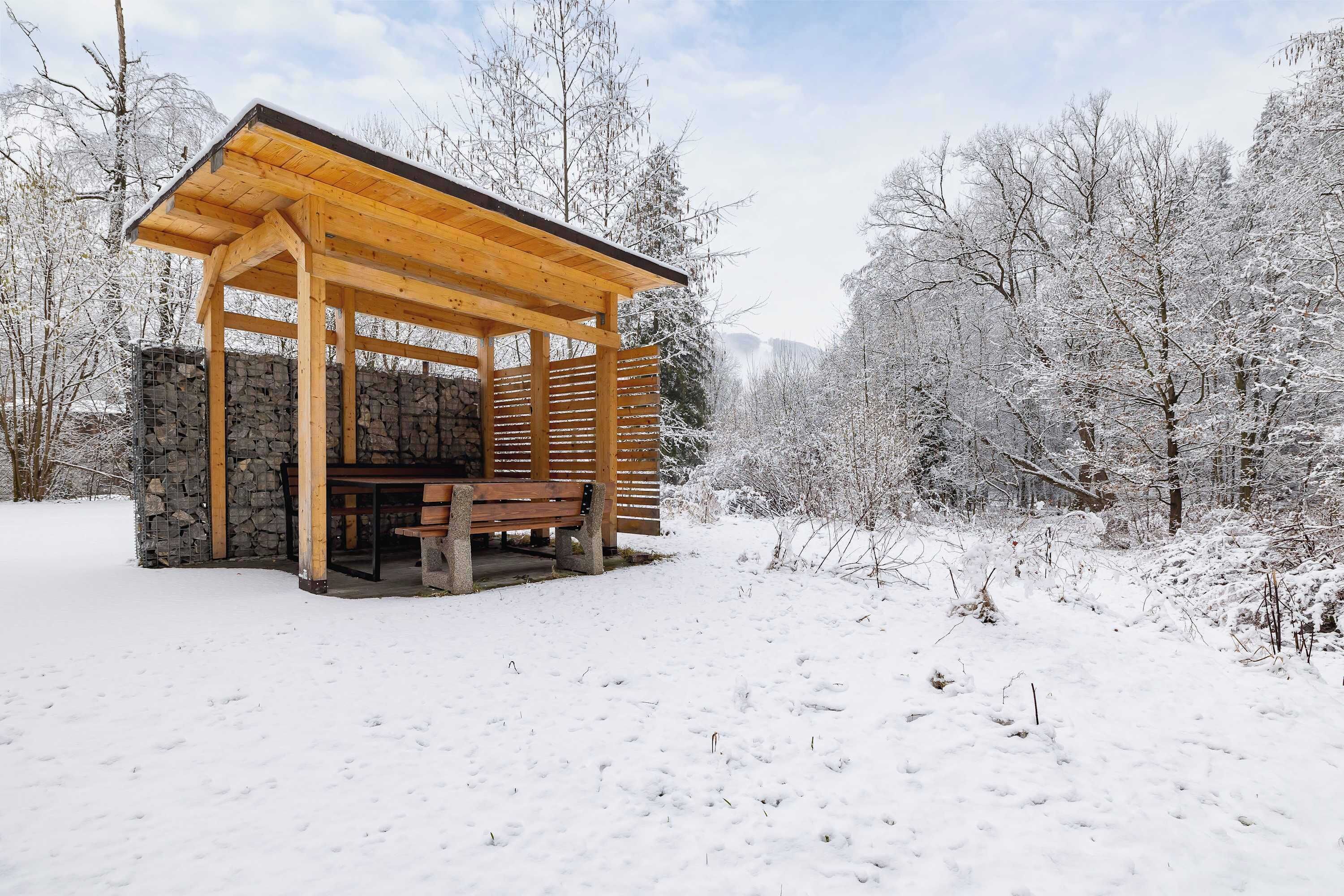 Un kiosque en bois avec un mur de pierre, un banc et une forêt enneigée en arrière-plan.