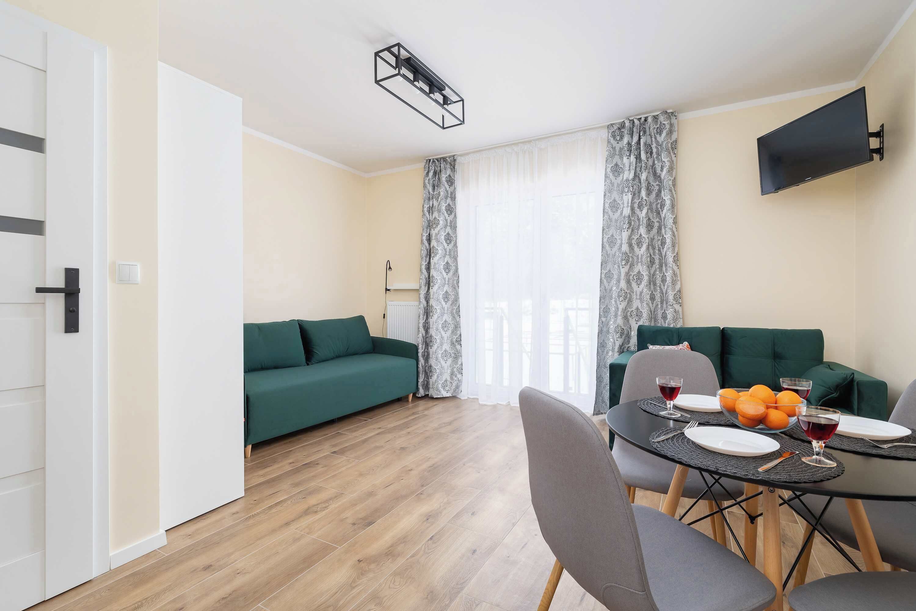 A modern living room with a dining area, green sofas, a flat-screen TV, and a wooden floor.