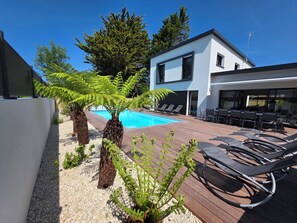 Un espace piscine extérieur moderne, doté de chaises longues, d’une terrasse en bois et d’un bâtiment blanc aux grandes fenêtres.