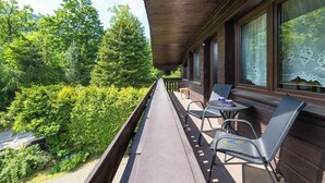 Ein Balkon mit Blick auf einen Wald, ein Tisch mit Stühlen und ein Fenster mit Spitzenvorhängen.