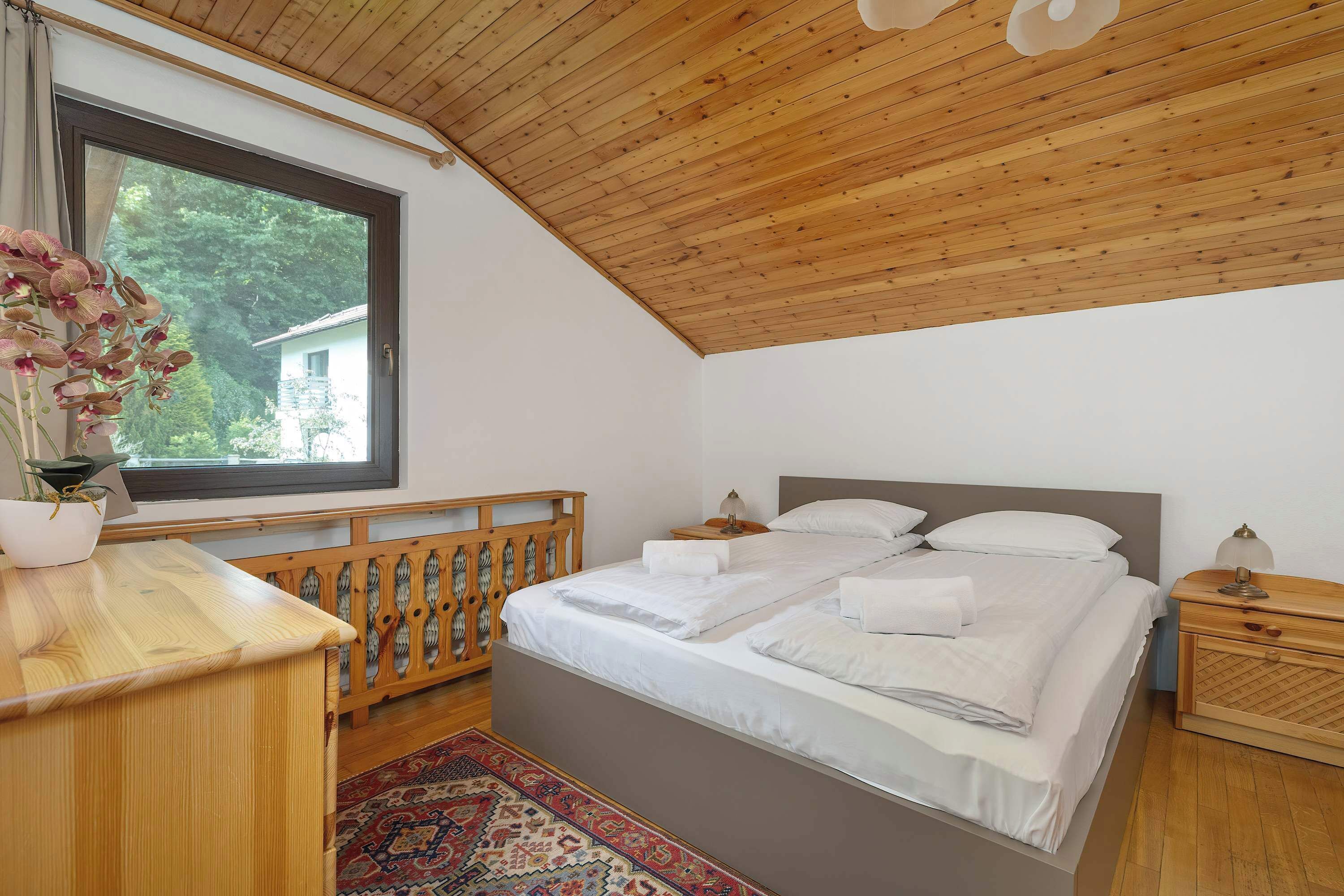 A bedroom with a wooden ceiling, a large bed with white linens, a wooden nightstand, and a window with a view of greenery.