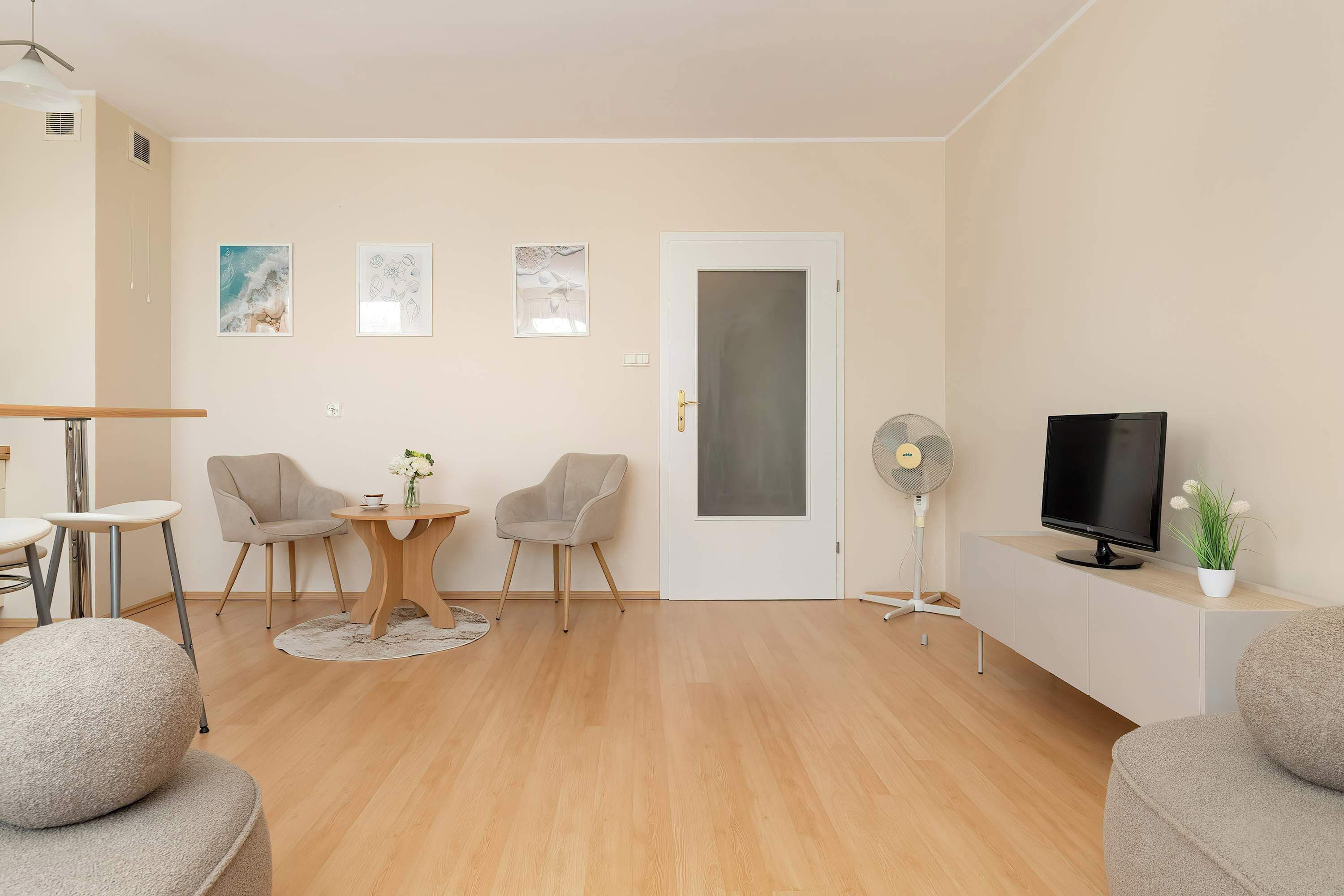 Un salon moderne avec un plancher en bois, deux fauteuils gris, une table ronde ornée d’un vase, un téléviseur posé sur un meuble blanc et une porte dotée d’un panneau en verre dépoli.