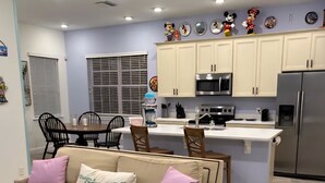 A modern kitchen with white cabinets, a stainless steel refrigerator, and a dining area with a beige sofa and a coffee machine.