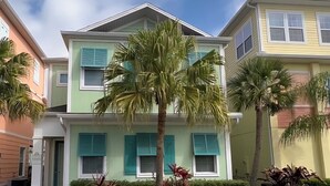 A two-story building with a green facade and teal shutters, surrounded by well-maintained landscaping including palm trees and shrubs.