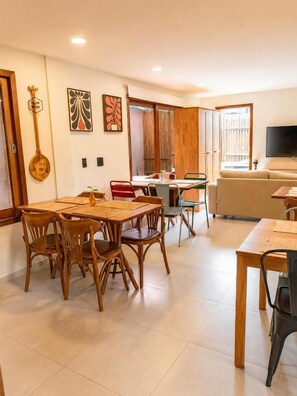 A dining area with a wooden table and chairs, a kitchen area with a counter and cabinets, and a living area with a sofa and television. - Pousada Divino Milagre (Passo de Camaragibe)