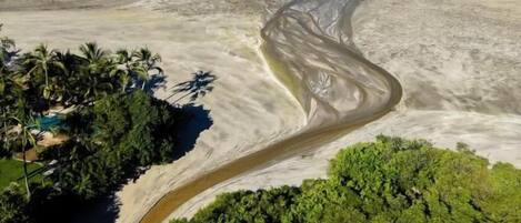 An aerial view of a beach with a winding river, dense greenery, and a clear blue sky.