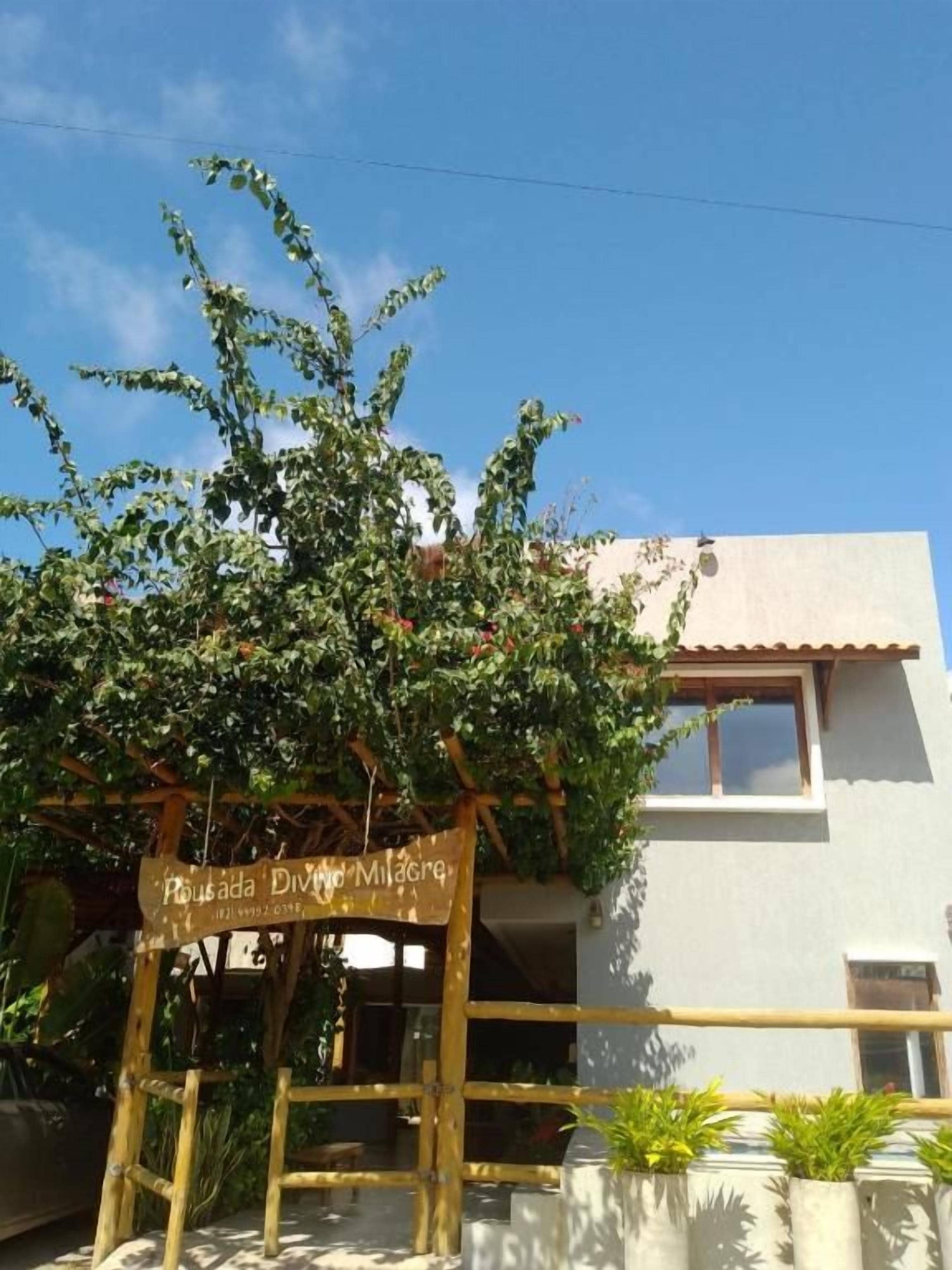 Um edifício com uma placa indicando 'Pousada Divino Milagre' e uma pérgola de madeira com plantas trepadoras.