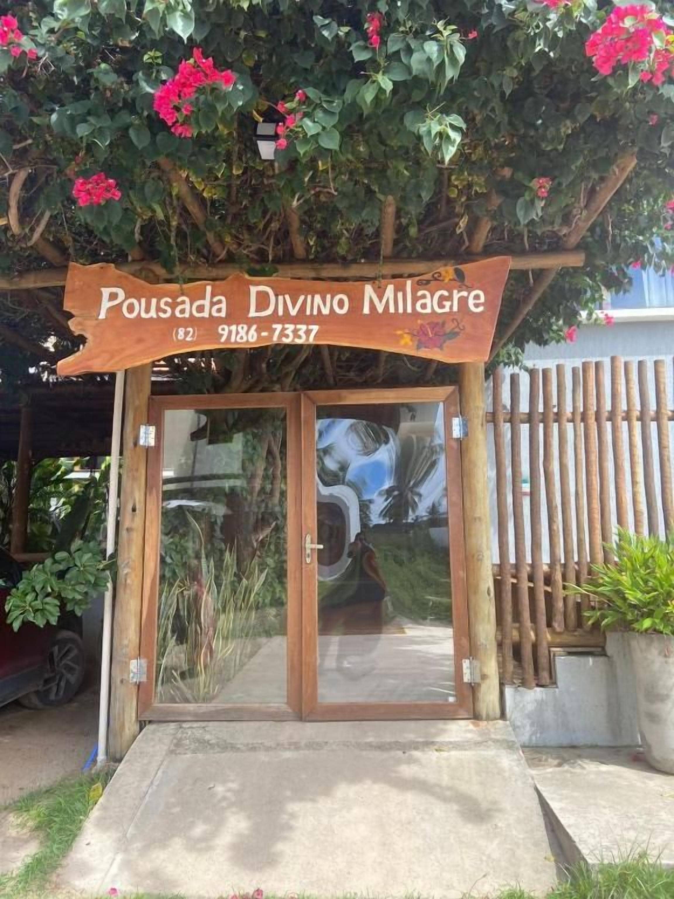 Uma entrada de madeira com porta de vidro, placa indicando 'Pousada Divino Milagre' e número de contato (82) 9186-7337.