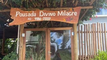 Uma entrada de madeira com porta de vidro, placa indicando 'Pousada Divino Milagre' e número de contato (82) 9186-7337.