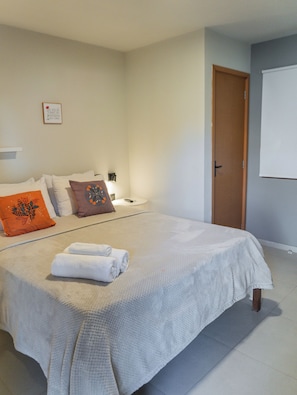 Una cama bien hecha con sábanas blancas, una almohada marrón y una almohada naranja. Hay una mesita con una lámpara y un marco de fotos en la pared.