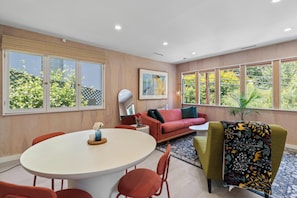 Una sala moderna con una mesa de comedor redonda, sillas rojas y verdes, un sofá rojo y un ventanal con vistas a árboles.
