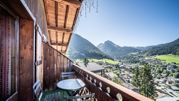 Een houten balkon met een tafel en stoelen, met uitzicht op een bergachtig landschap en een dorp beneden.