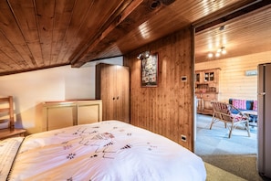 A wooden interior with a bed, a wooden wardrobe, and a dining area with a chair and table.