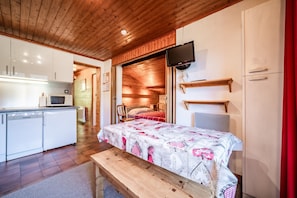 Ein gemütlicher Raum mit Bett, einer kleinen Küchenzeile, einem Fernseher und einer Holzbalkendecke.