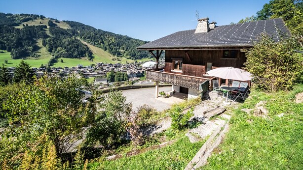 Een houten chalet met balkon, omgeven door groen en uitzicht op een dorp en heuvels.