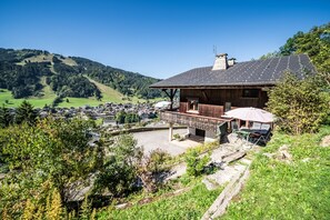 Een houten chalet met balkon, omgeven door groen en uitzicht op een dorp en heuvels.