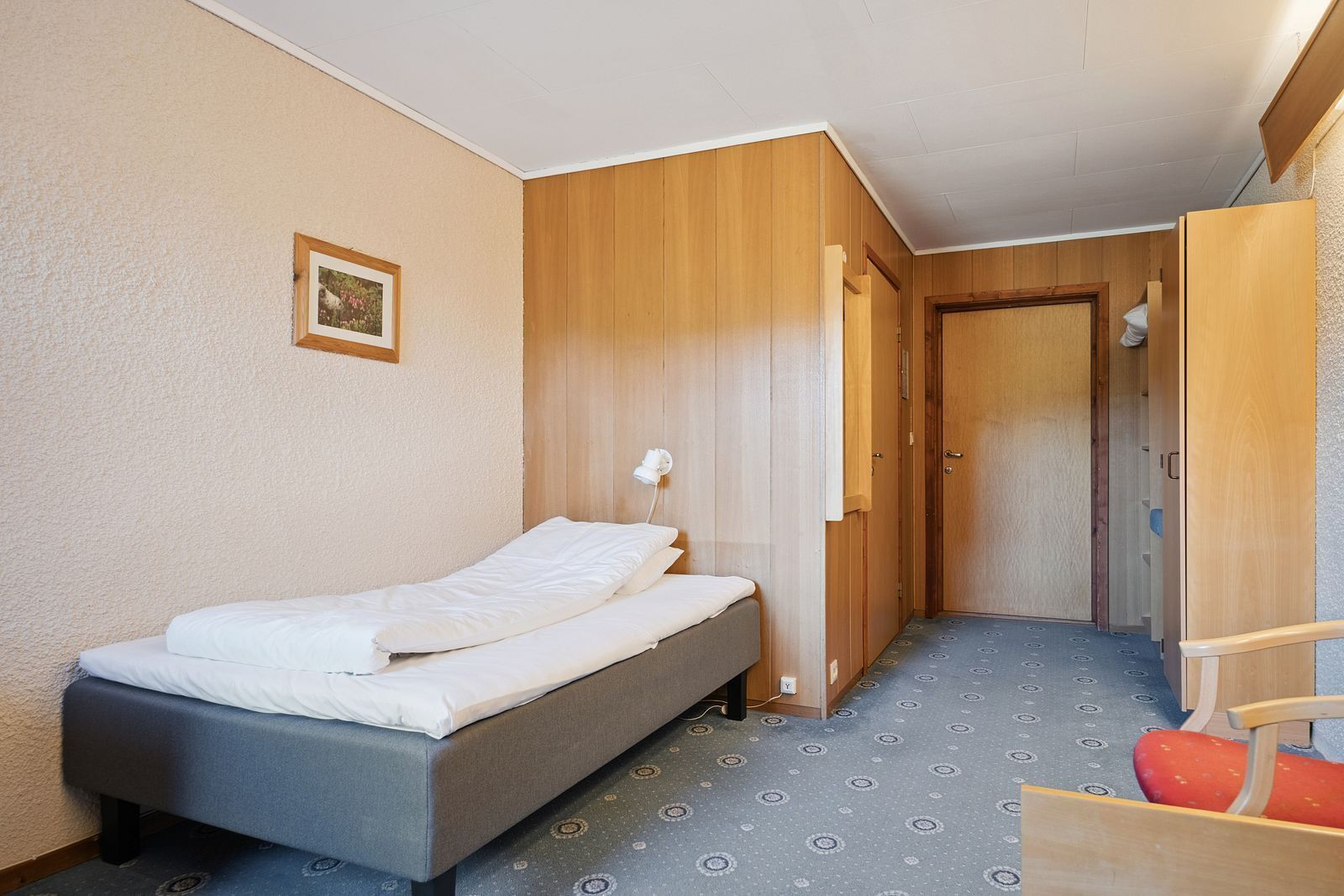 A small, single-bed room with a wooden wardrobe, a bedside lamp, and a patterned carpet.