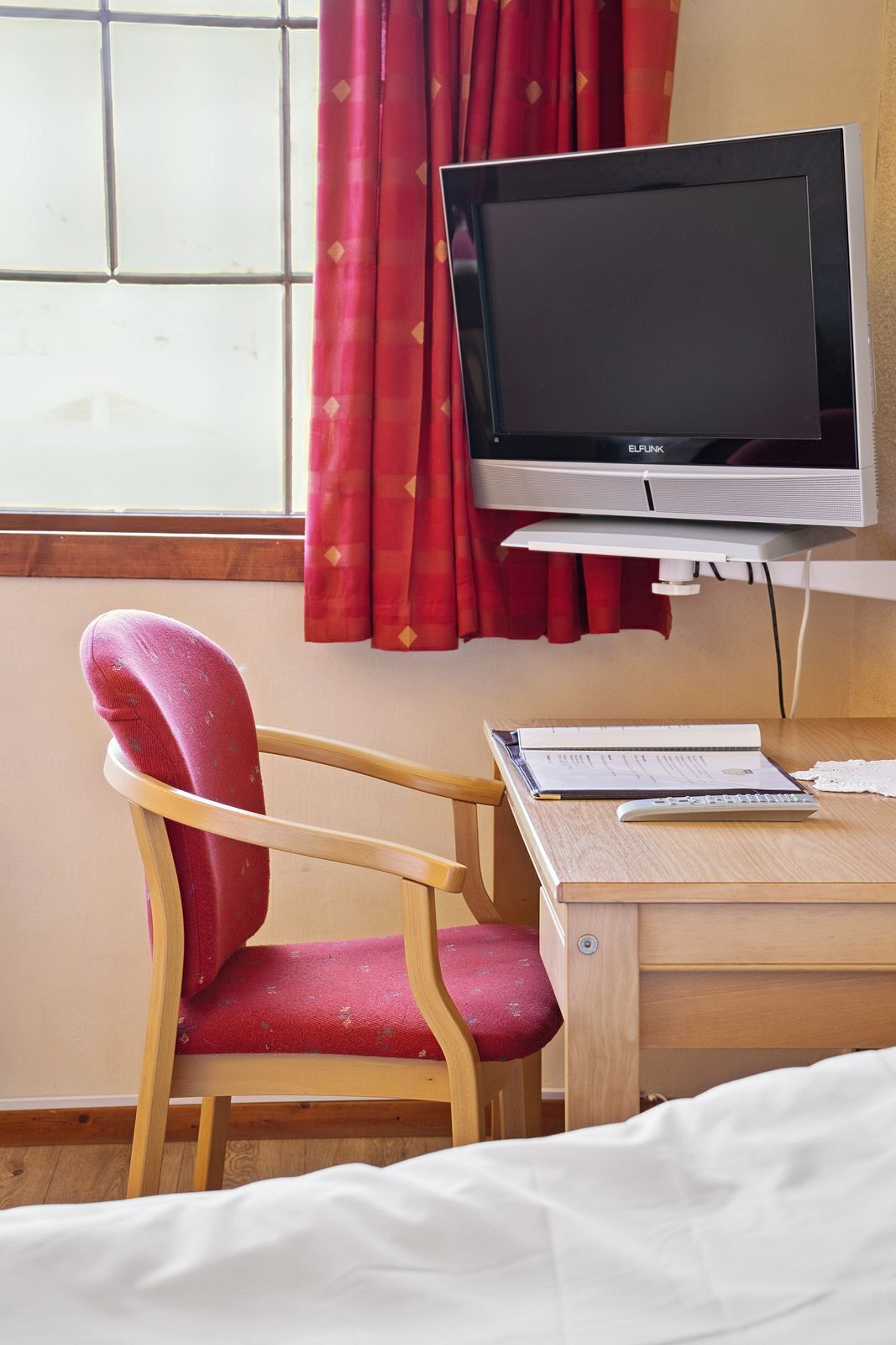 A hotel room with a wooden desk, a red chair, a bed, and a television on a stand.