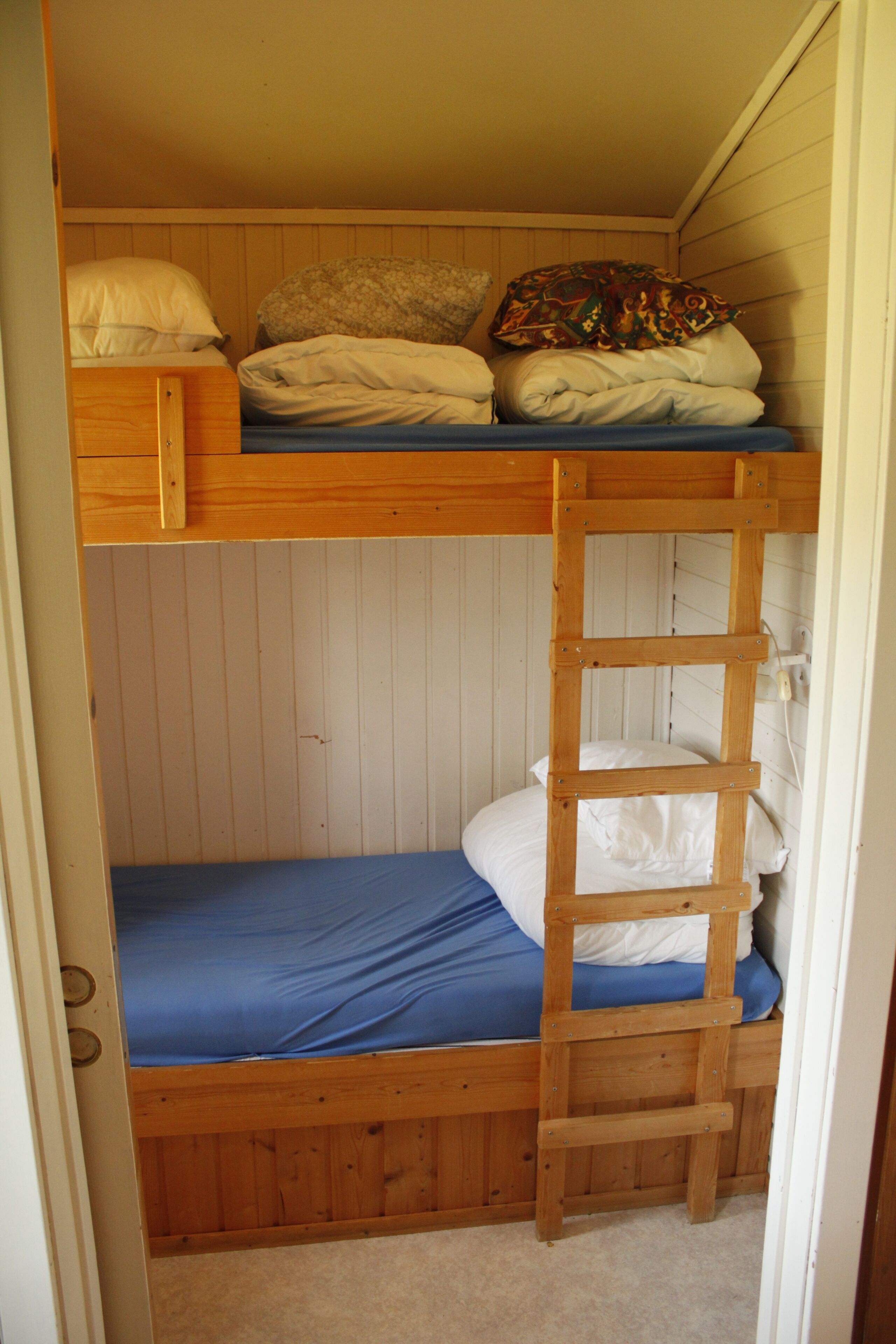 Una cama litera con escalera de madera, colchón azul y almohada blanca.