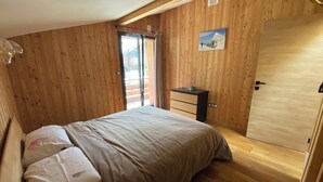 A bedroom with wooden walls, a bed with a beige blanket, a small dresser, and a door. - Hideaway at the foot of Mont Blanc (Les Houches)