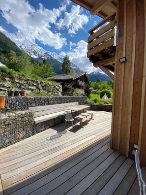 Una terraza de madera con mobiliario exterior, un muro de piedra y montañas al fondo.