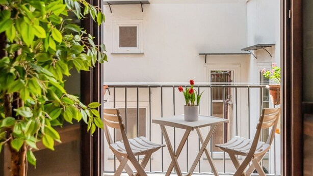 Un balcón con mesa y sillas, una maceta con flores rojas y vista del exterior del edificio.