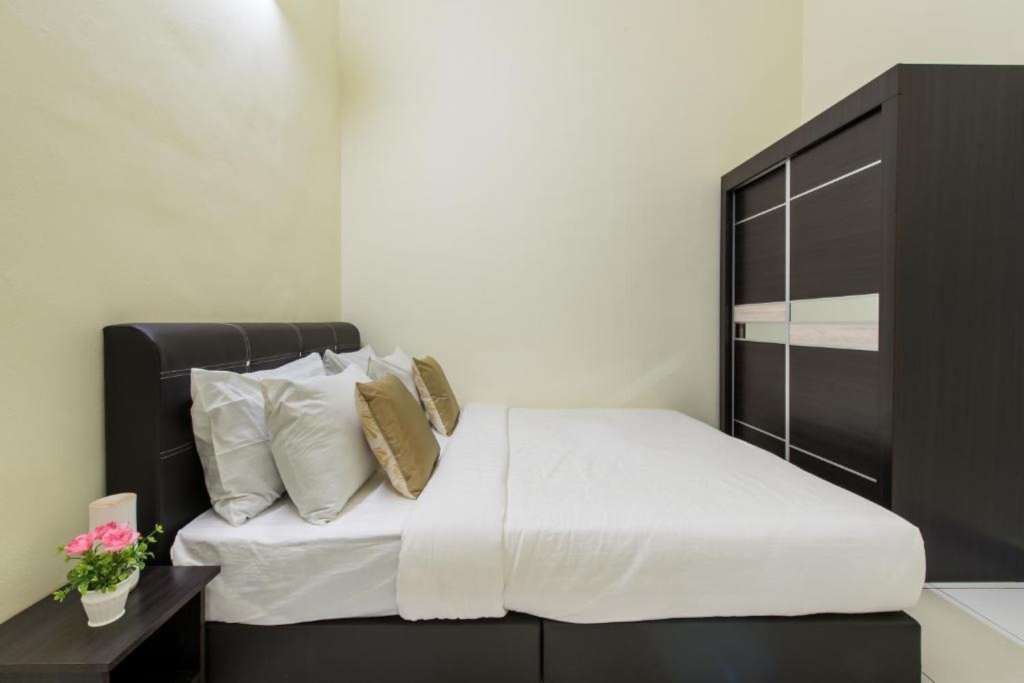 Een bed met witte beddengoed en meerdere kussens, een donker hoofdbord, een nachtkastje met een plant, en een donkere kast met een lichtpaneel.