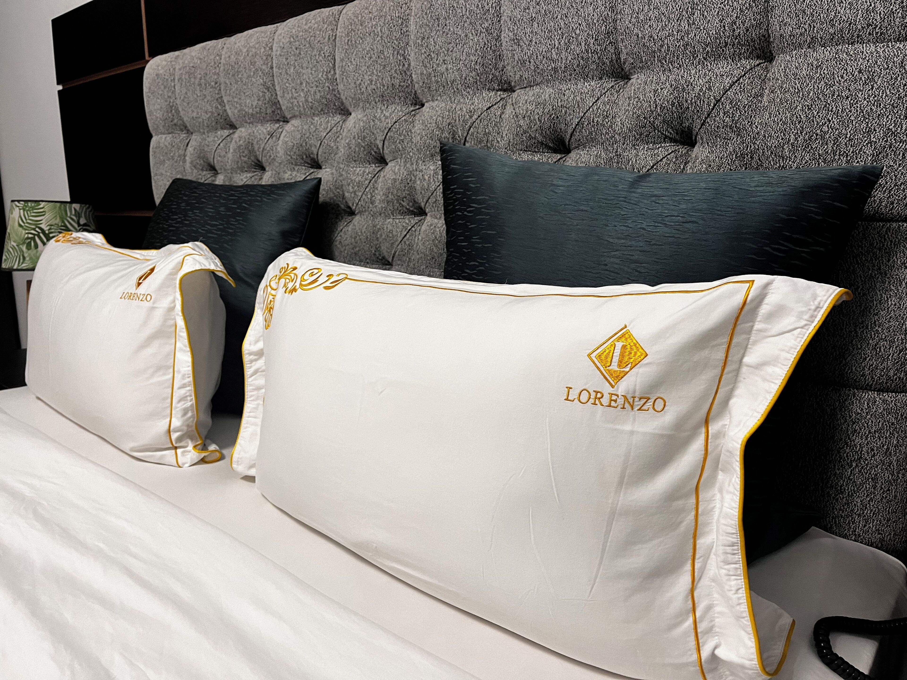 A bed with white bedding and pillows featuring a gold embroidered logo.