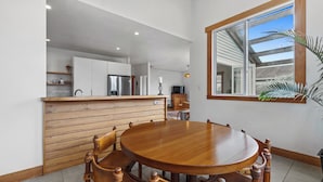 A dining area with a wooden table and chairs, a kitchen in the background, and a large window with a view of the outside.